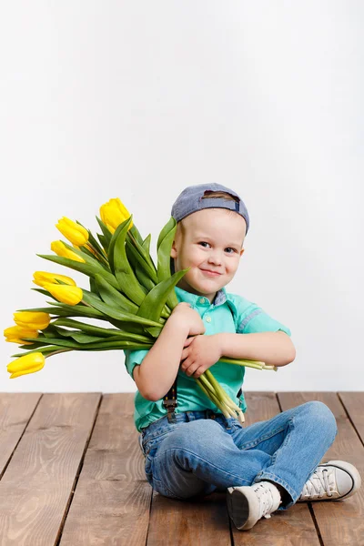 Smiling boy holding a bouquet of yellow tulips in hands sitting on wooden floor — Stock Photo, Image