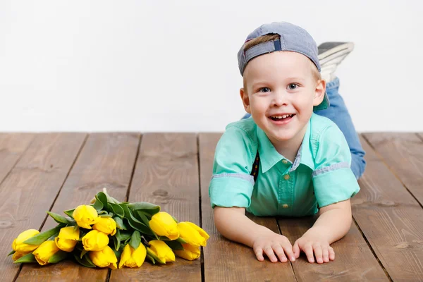 Smiling boy holding a bouquet of yellow tulips in hands sitting on wooden floor — Stock Photo, Image