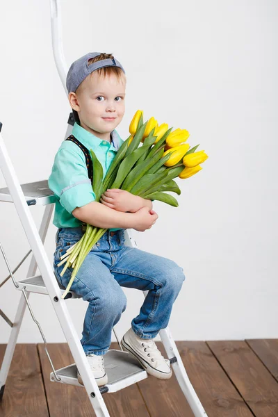 Smiling boy holding a bouquet of yellow tulips in hands sitting on wooden floor — Stock Photo, Image