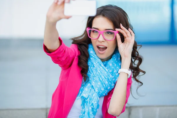 Happy young girl making funny face while taking pictures of herself through cellphone, over white background — Stock Photo, Image