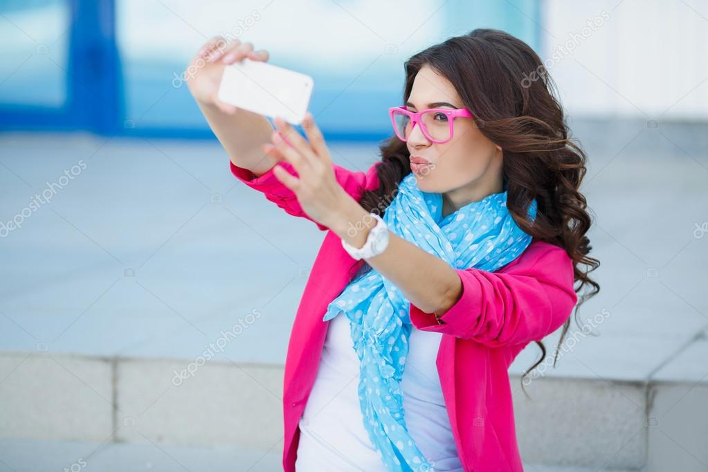 Happy young girl making funny face while taking pictures of herself through cellphone, over white background