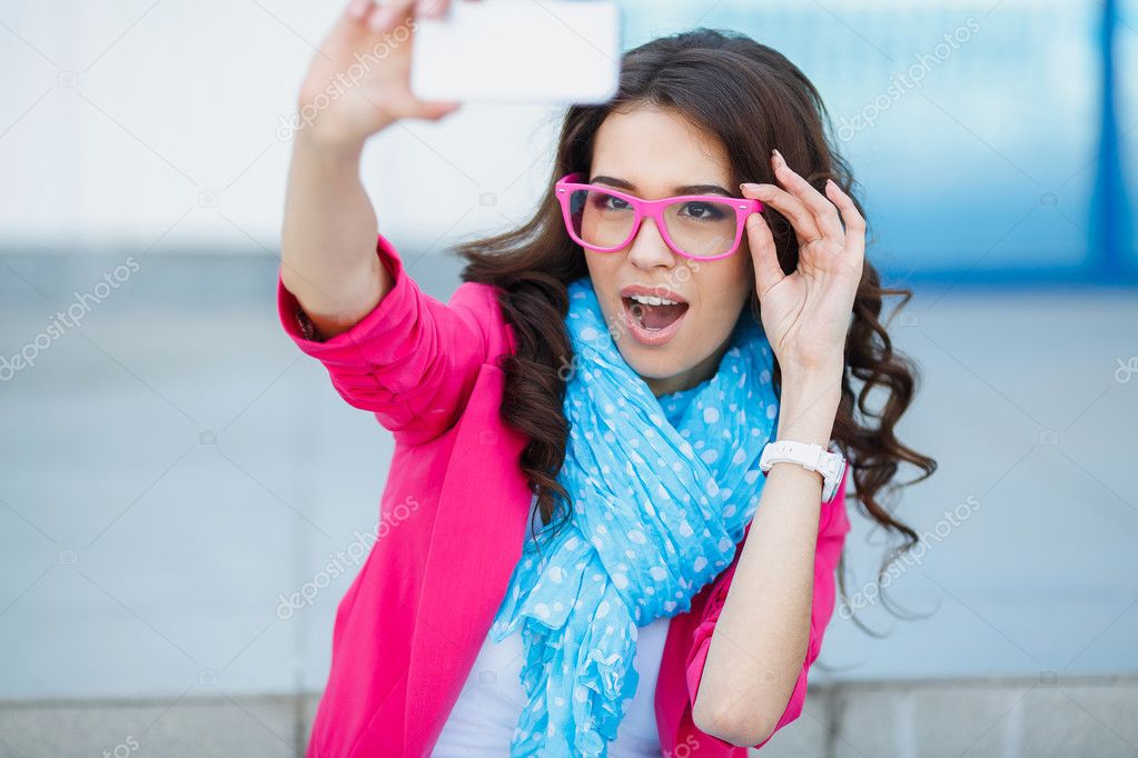Happy young girl making funny face while taking pictures of herself through cellphone, over white background
