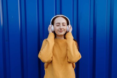 Duygu yüklü, kızıl saçlı genç bir kadın. Açık havada kulaklıkla müzik dinliyor. 