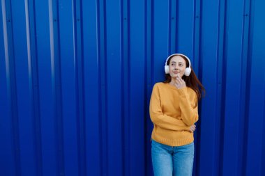 Duygu yüklü, kızıl saçlı genç bir kadın. Açık havada kulaklıkla müzik dinliyor. 