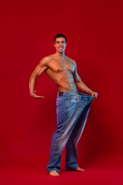 Genç adam büyük kotlarla ne kadar kilo verdiğini gösteriyor. Konsepte uygun yağ. Kırmızı arkaplanda fitness posteri