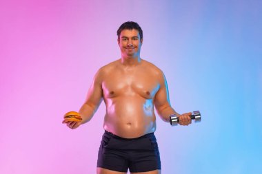 Şişman adam hamburgerle halterli fitness arasında seçim yapıyor. Pembe arka planda izole edilmiş hamburger yiyen sporcular değil..