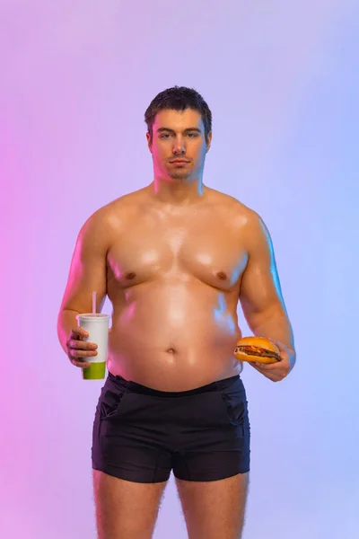 Fat man with burger and soda. Not sporty men drinking and eating hamburger isolated on pink background. — Stockfoto
