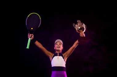 Tennis player with cup and racquet in fire celebrating a flawless victory in the tournament. Beautiful girl teenager and athlete with racket in pink sporswear and hat on tennis court.