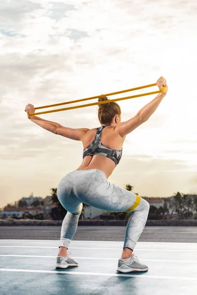 Allenamento con le fasce di fitness. Donna sportiva con corpo abbronzato sottile in abbigliamento sportivo in posa su sfondo cielo. — Foto Stock