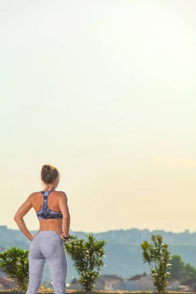 Donna sportiva con corpo in forma in abbigliamento sportivo in posa su sfondo cielo su pista da corsa. Vista posteriore verticale. — Foto Stock