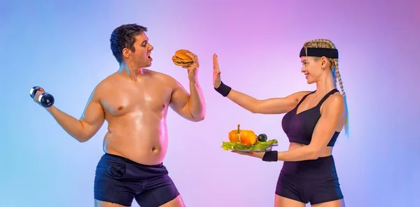A kövér ember választja a hamburgert és a súlyzóst. Az edző lány azt mondja neki, hogy hagyja abba, és zöldségeket ad a gyorskaja helyett. Nem sportos férfiak eszik hamburger elszigetelt rózsaszín alapon. Stock Fotó