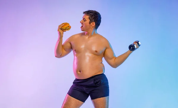 Fat man with burger and dumbbell. Not sporty men eating hamburger isolated on pink background