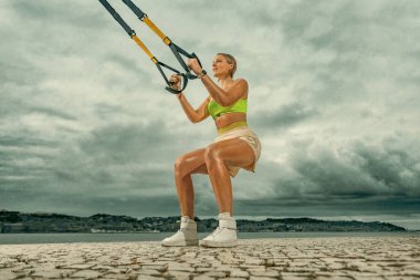 Açık havada trx fitness kayışlarıyla şınav çeken bir kadın. Spor kıyafetli, zinde bir kadın gökyüzünde poz veriyor.