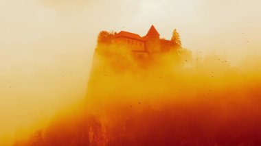 Lake Bled castle in orange clouds. Dramatic view on Blejski grad. Foggy castle on mountain with birds flying by. Handheld shot. Lake Bled, Slovenia.