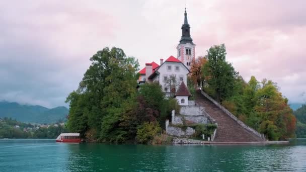スロベニアではボートから湖のせせらぎ 雪に覆われたジュリアン アルパイン山脈に対するマリア教会の仮定の日没の眺め 湖の水の中に反映教会 教会の周りを回転する手持ち撮影 — ストック動画