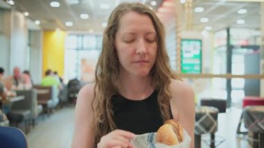 Woman with burger in a fast food restaurant. Woman opening wrapping paper, holding, biting the burger and savouring food. Fast snack on the go