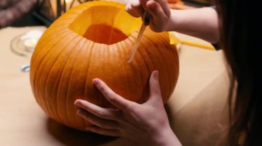 Preparing pumpkin for Halloween. Woman sitting and carving with knife face details of halloween Jack O Lantern pumpkin at home for her family.