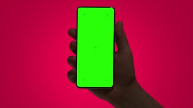 Hand holding mobile phone. Dark hand silhouette holds smartphone against red background. Green chromakey screen with markers. Hand lifting phone up and holding it still.