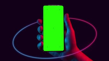 Mobile phone in hand. Holding smartphone with red blue neon circle on dark background. Green chromakey screen with markers. Hand lifts phone up and holds it still.