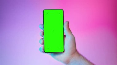 Mobile phone in hand. Holding smartphone with colorful red blue background. Green chromakey screen with markers. Neon lighting. Hand lifts phone up and holds it still.