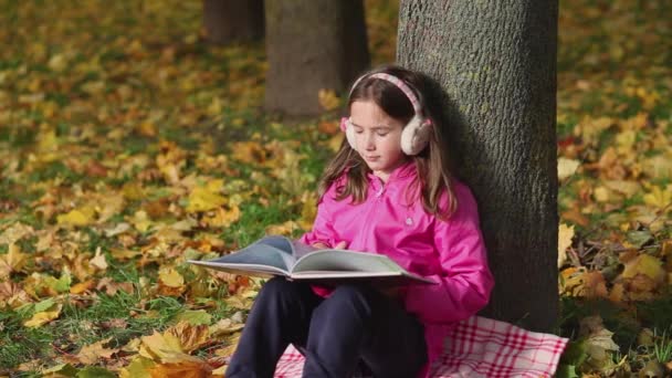 Linda niña lee un libro sentado debajo de un árbol. Hojas coloridas caen del árbol. — Vídeo de stock