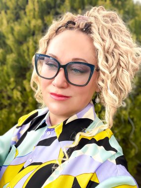 Gözlüklü, kıvırcık sarı saçlı ve yeşil thuja arka planında renkli bluzu olan güzel bir kadının portresi.