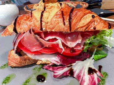 Fresh croissant sandwich with dried ham on a bed of tomatoes and lettuce with pesto and balsamic sauce on breakfast closeup