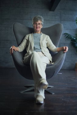Sophisticated elegant beautiful 50s attractive middle aged smiling woman model sitting in chair looking at camera at home