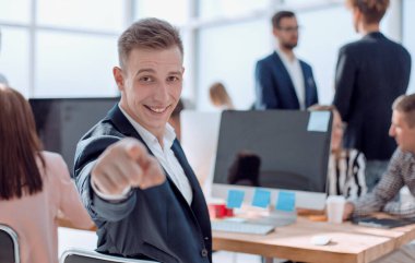 Gülümseyen genç bir çalışan masasında oturuyor ve seni işaret ediyor.