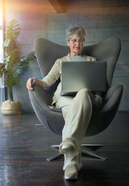Mujer sentada en silla moderna cerca de la ventana en la habitación acogedora luz en casa. Ella está trabajando en el ordenador portátil en un ambiente relajante — Foto de Stock