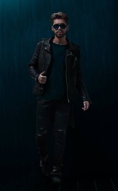 Deri ceketli, kot pantolonlu ve botlu sıradan bir adamın stüdyo duvarına yaslanmış tüm vücudu.