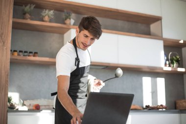 Çekici genç adam yemek pişirirken dizüstü bilgisayara bakıyor.