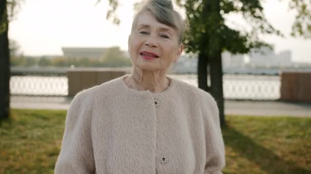 Egy elegáns, idős nő mosolygó portréja, ahogy a kamerát nézi a szabadban a modern városi utcában. — Stock videók