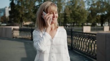 Modern şehirde yaz günü cep telefonuyla konuşan neşeli yetişkin bir kadın.