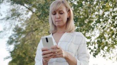 Açık havada gülümseyen akıllı telefon kullanan güzel sarışın bir kadının portresi.