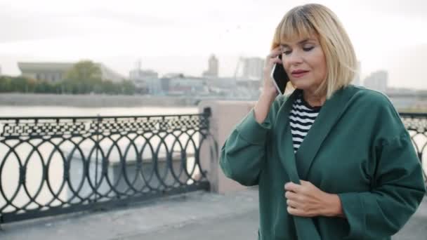 Beautiful woman speaking on mobile phone walking outdoors along river embankment — Stock Video