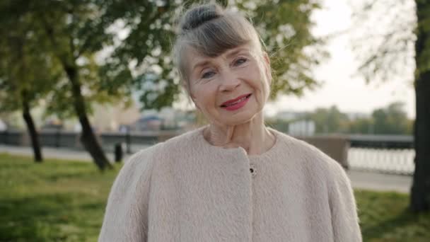 Медленное движение положительной пожилой женщины улыбаясь и показывая ОК жеста руки стоя снаружи — стоковое видео