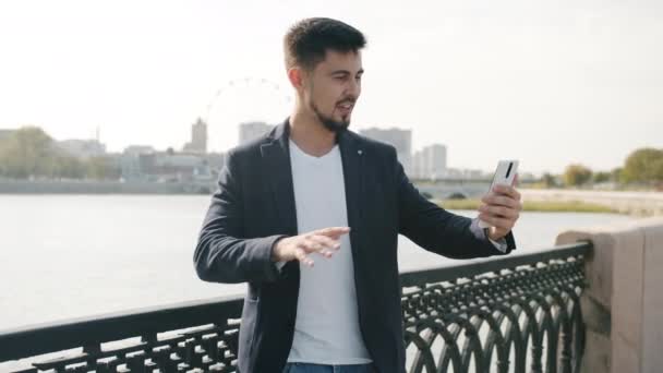 Geselliger junger Mann spricht und gestikuliert mit dem Zeigefinger auf den Fluss und macht im Sommer Online-Videoanrufe im Freien — Stockvideo