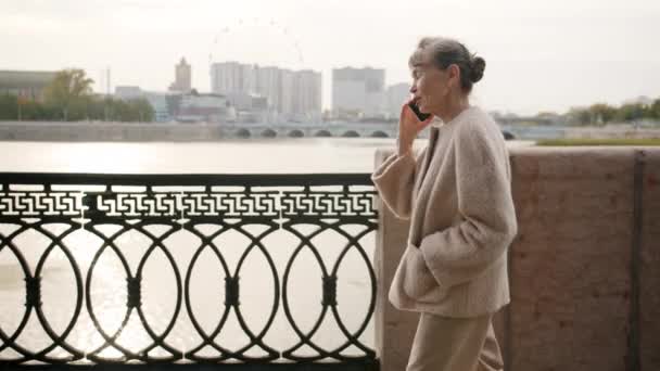 Вид сбоку пожилой дамы, гуляющей по мобильному телефону на городском пешеходном мосту в летний день — стоковое видео