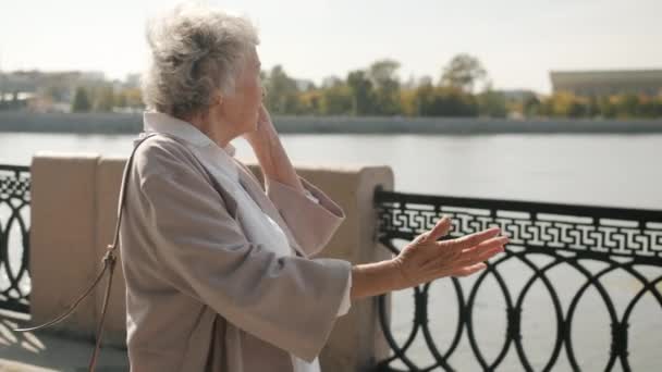 Glada äldre kvinna promenader längs floden vallen och chatta på mobiltelefon på sommardagen — Stockvideo
