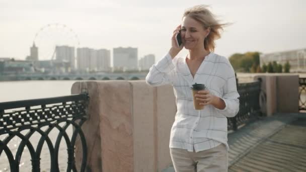 Cute blondynka czat na telefon komórkowy śmiech gospodarstwa iść kawa spacery na świeżym powietrzu w nabrzeżu rzeki — Wideo stockowe