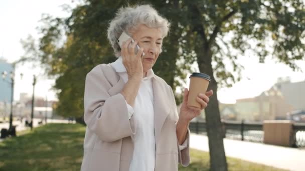 Frumoasă doamnă bătrână vorbind pe telefonul mobil și bea pentru a merge cafea în aer liber pe strada orașului — Videoclip de stoc