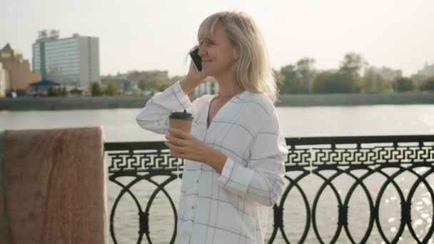 Wesoła blondynka rozmawiająca przez telefon komórkowy pijąca kawę spacerując wzdłuż nabrzeża rzeki w mieście — Wideo stockowe