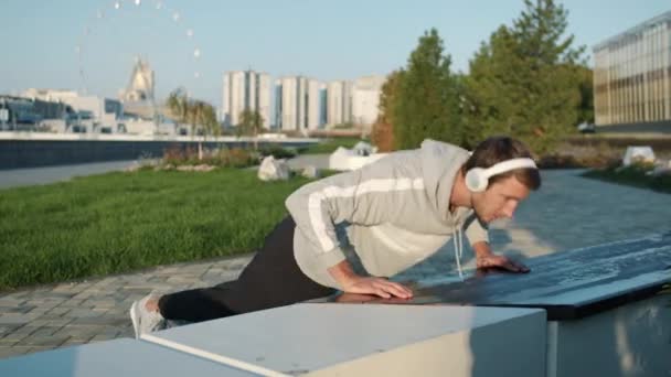 Portrét sportovního chlapa, který dělá kliky a poslouchá hudbu se sluchátky venku ve městě — Stock video