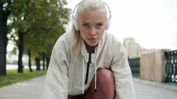 Poważna młoda blondynka w słuchawkach szykuje się do maratonu i biegnie — Wideo stockowe