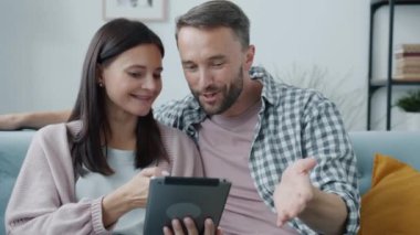 Tablet kullanan neşeli genç çift gülümseyerek ekranlara dokunuyor ve evde konuşuyorlar.