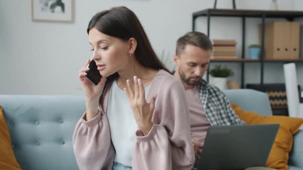 Boos jonge vrouw spreken op mobiele telefoon, terwijl de man werkt met laptop op de achtergrond — Stockvideo