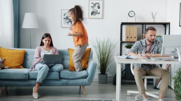 Los padres que trabajan con computadoras desde casa mientras la hija hiperactiva tonteando — Vídeo de stock