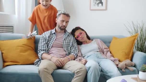 Utslitte foreldre som sitter på sofaen mens en liten jente løper rundt og skriker og hopper innendørs – stockvideo