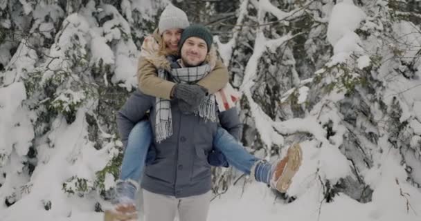 Медленный портрет милой пары, развлекающейся в зимнем лесу мужчина держит женщину в спине — стоковое видео